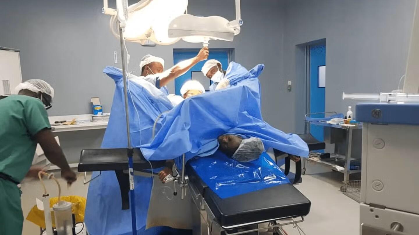 Une équipe chirurgicale composée de plusieurs médecins et infirmiers procède à une intervention de réparation de fistule obstétricale dans une salle d’opération stérile. Une patiente est allongée sur la table d’opération, entourée de champs opératoires bleus et de matériel médical Une équipe chirurgicale en pleine intervention de réparation de fistule obstétricale, dans le cadre de la campagne régionale 2025